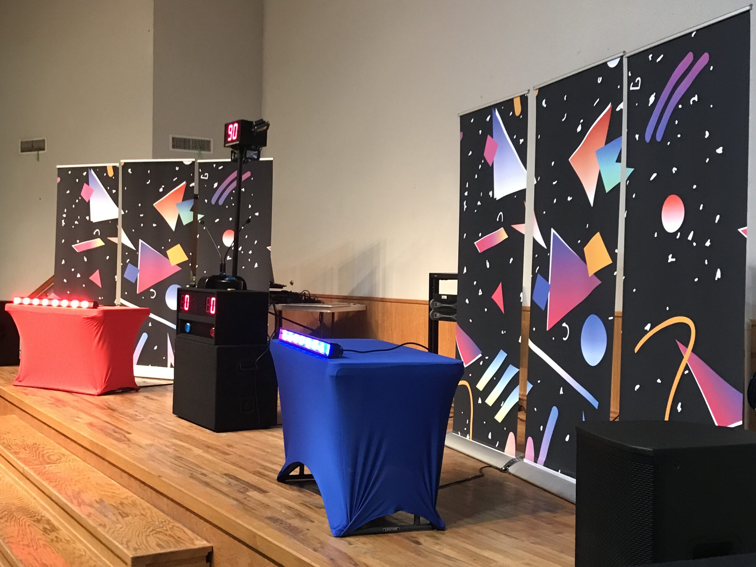 A colorful quiz setup features red and blue tables with scoreboards, flanked by vibrant backdrop panels, in a dynamic event space.