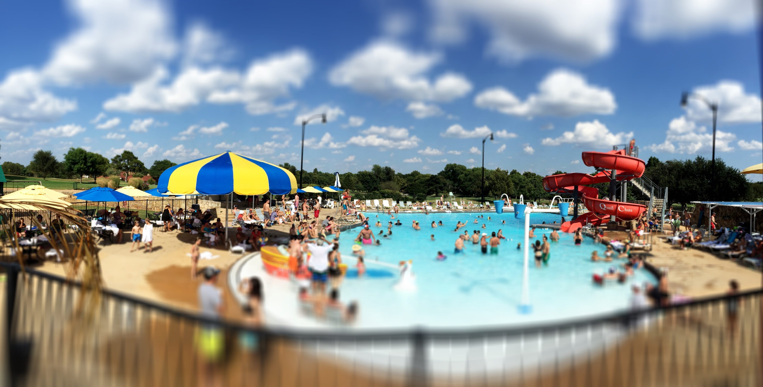 Crowded outdoor pool area with colorful umbrellas, a water slide, and families enjoying the sun. The scene captures a lively summer atmosphere.