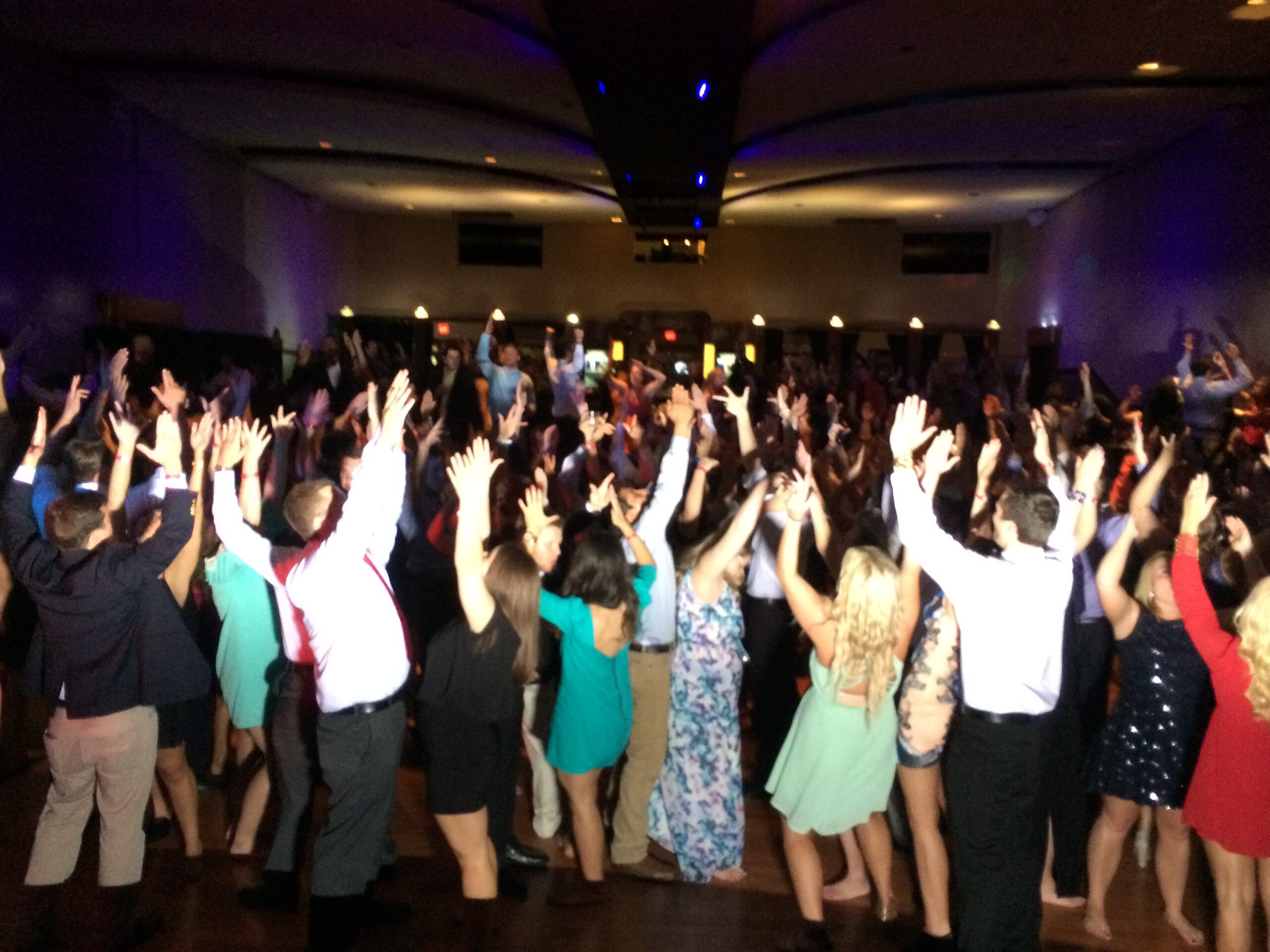 A lively crowd dances with hands raised, celebrating an event in a dimly lit venue. The vibrant atmosphere reflects joy and camaraderie.