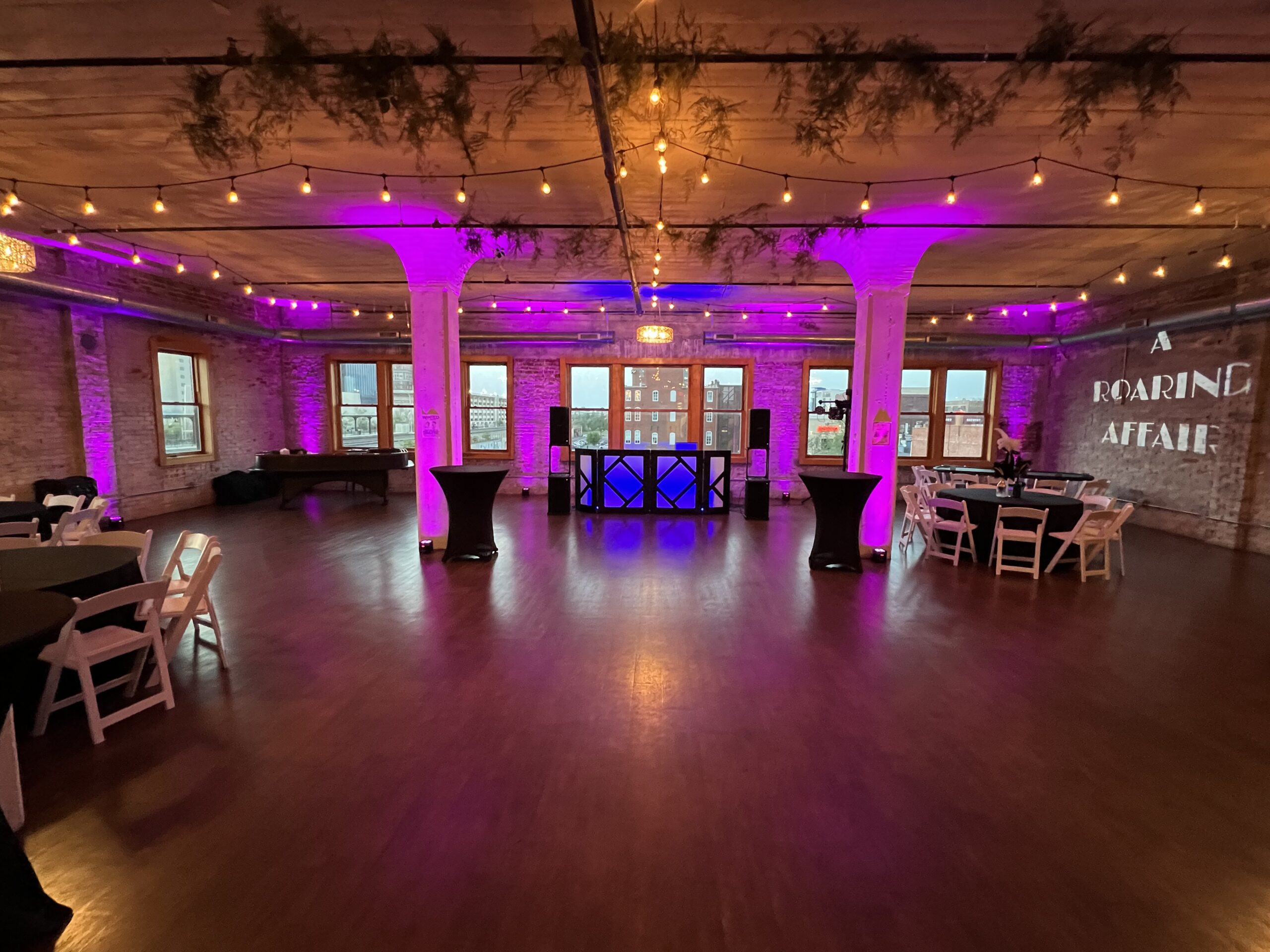 Vibrant event space with purple lighting, tables set for guests, and a DJ booth, showcasing a "Roaring Affair" theme against a brick backdrop.