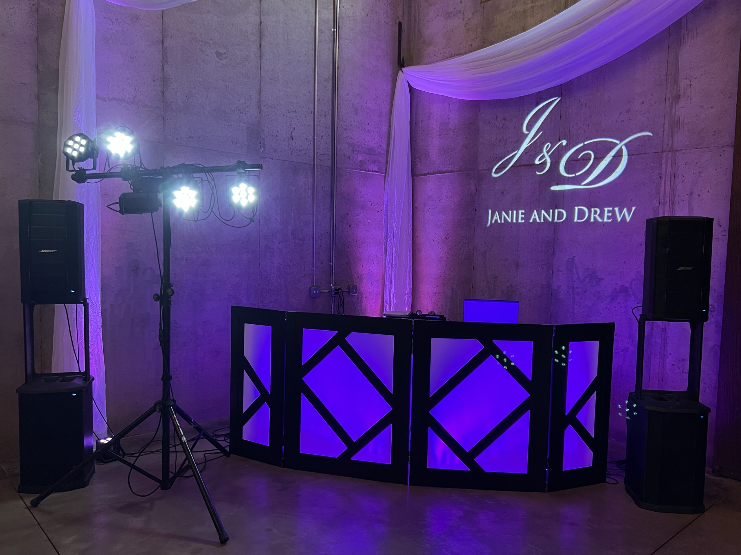 DJ setup featuring purple lighting and speakers, with a personalized projection reading "J & D Janie and Drew" against a concrete wall, ideal for a wedding or event.