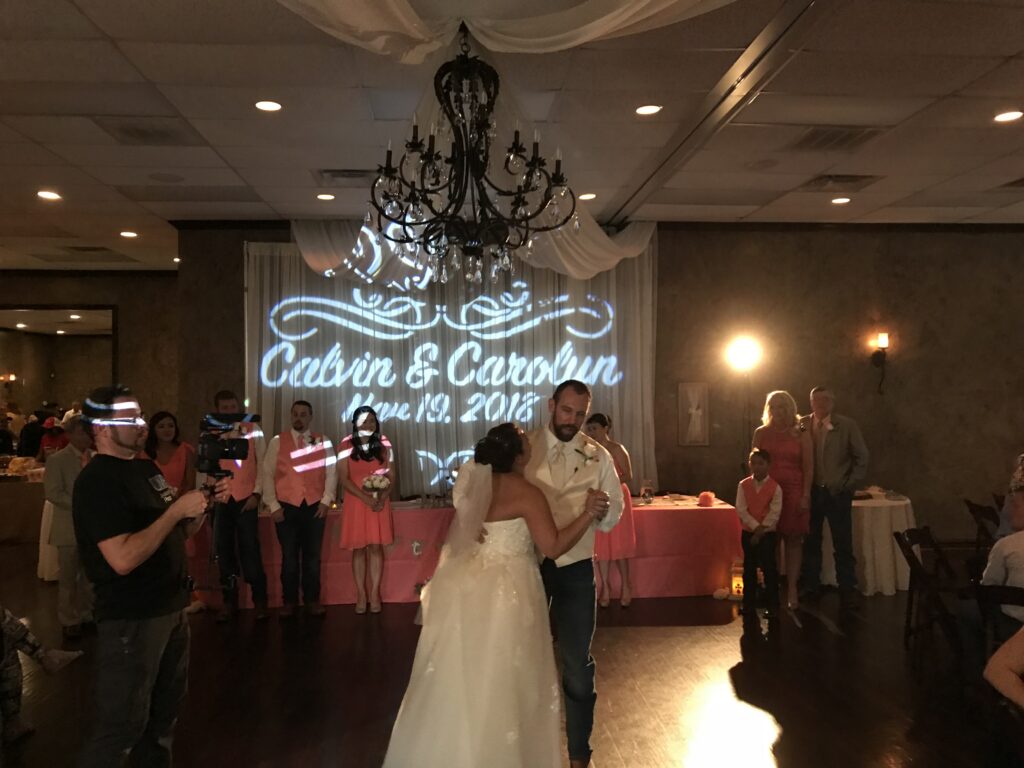 A bride and groom dance at their wedding reception, with guests in coral attire watching. The event features a light projection of their names and date.