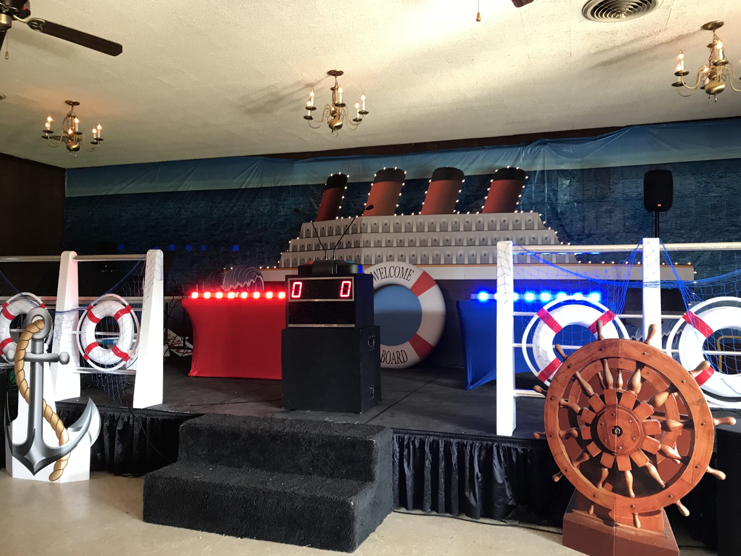 Nautical-themed stage setup featuring a large ship backdrop, lifebuoys, an anchor, a ship's wheel, and illuminated scoreboards, suitable for a maritime event.